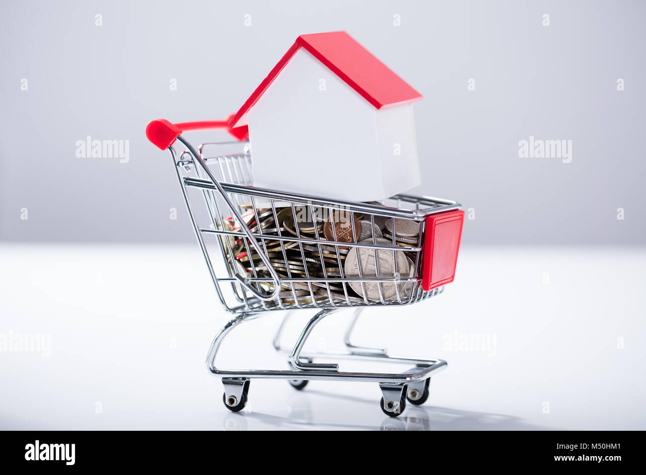 Cumulo di monete e modello di casa all'interno della miniatura carrello di shopping su sfondo bianco Foto Stock