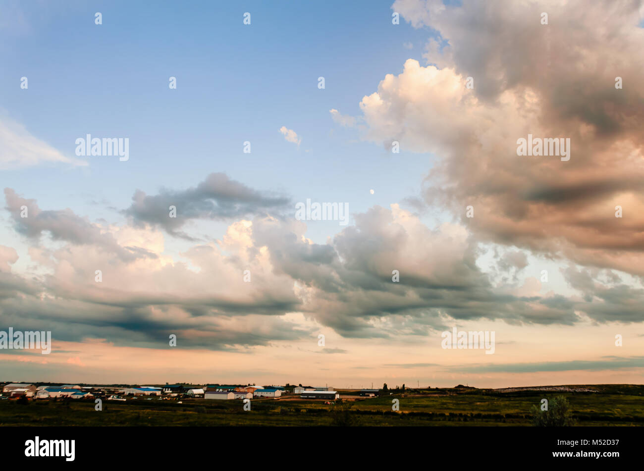 Arancione e bianco delle nuvole, evidenziata dalla Rising Sun, contro il cielo blu, edifici agricoli e cespugli verdi in background Foto Stock