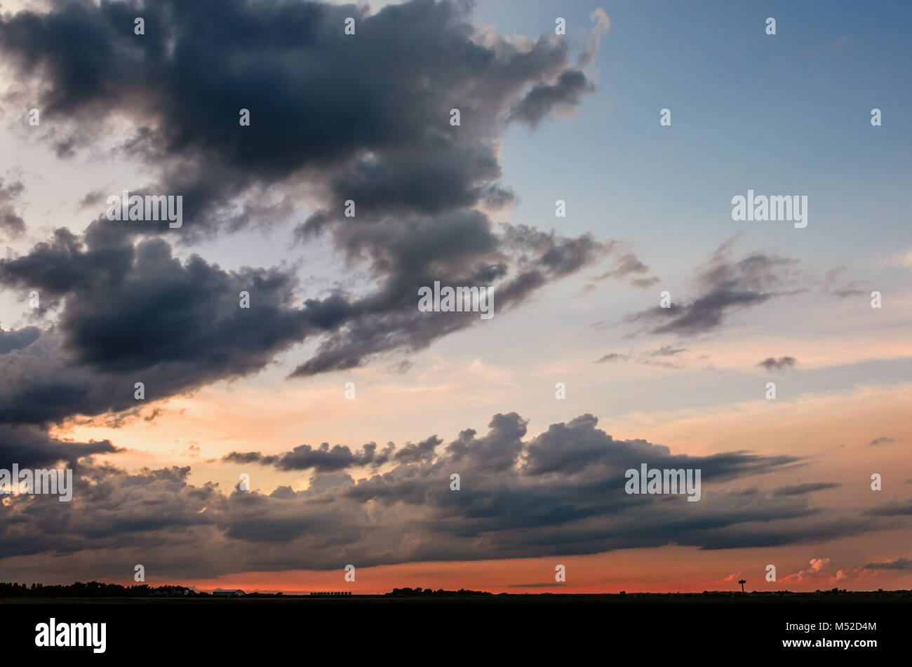 Nuvole grigie e una striscia scarlatta del tramonto su un campo di fattoria con fienili, contro un cielo blu, edifici agricoli e gli alberi in background Foto Stock