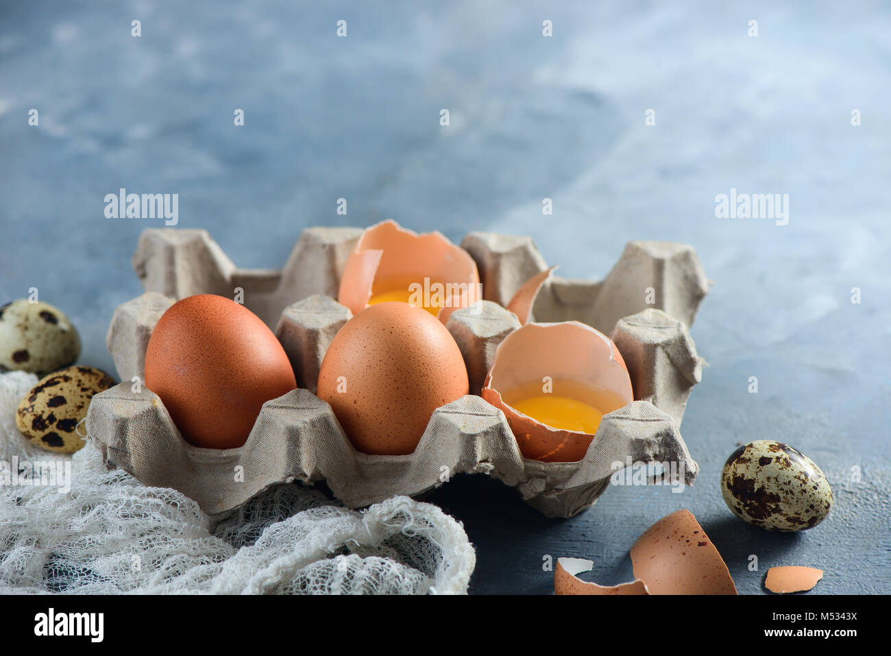 Quaglie fresche e uova di gallina, intere e rotte, in un imballo di carta su un sfondo di calcestruzzo. Materie prime per la Pasqua di cottura. Close-up con copia Foto Stock