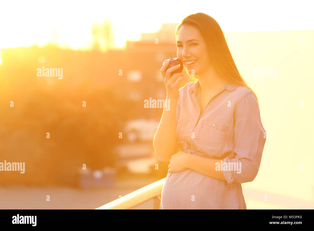 Donna incinta in possesso di un apple e guardando a voi in un balcone al tramonto Foto Stock