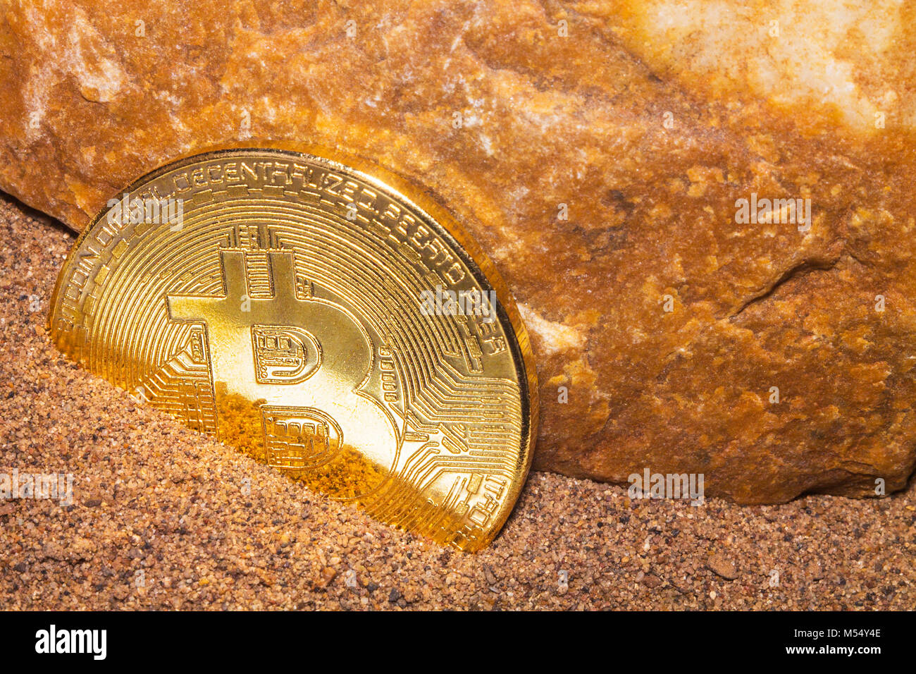 La moneta in oro della moneta crypto Bitcoin risiede nella sabbia vicino alla roccia di colore arancione. Foto Stock