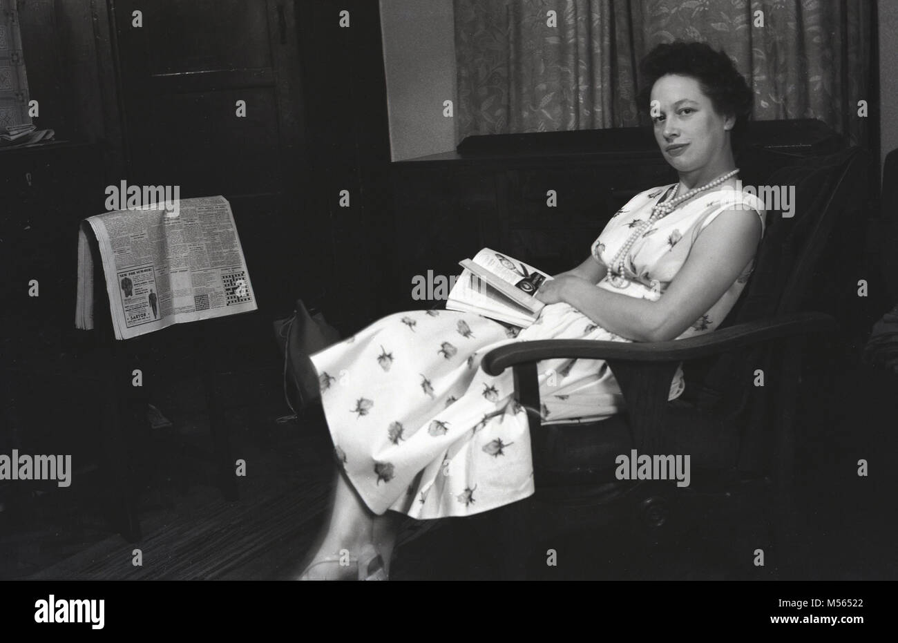 1940s, Manchester, Inghilterra, foto storiche che mostra una donna che indossa un abito lungo e una collana di perle seduto in una sedia nella sua camera anteriore con un libro in grembo e una copia del Manchester Guardian dalla credenza. Foto Stock
