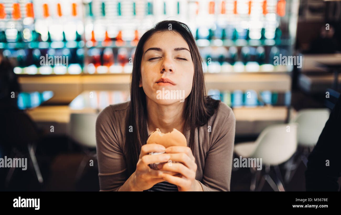 Giovane donna mangiare hamburger grassi.Craving fast food.godendo colpevole piacere,mangiare cibo spazzatura.soddisfatto l'espressione.rompendo la dieta regole,rinunciare a dieta Foto Stock