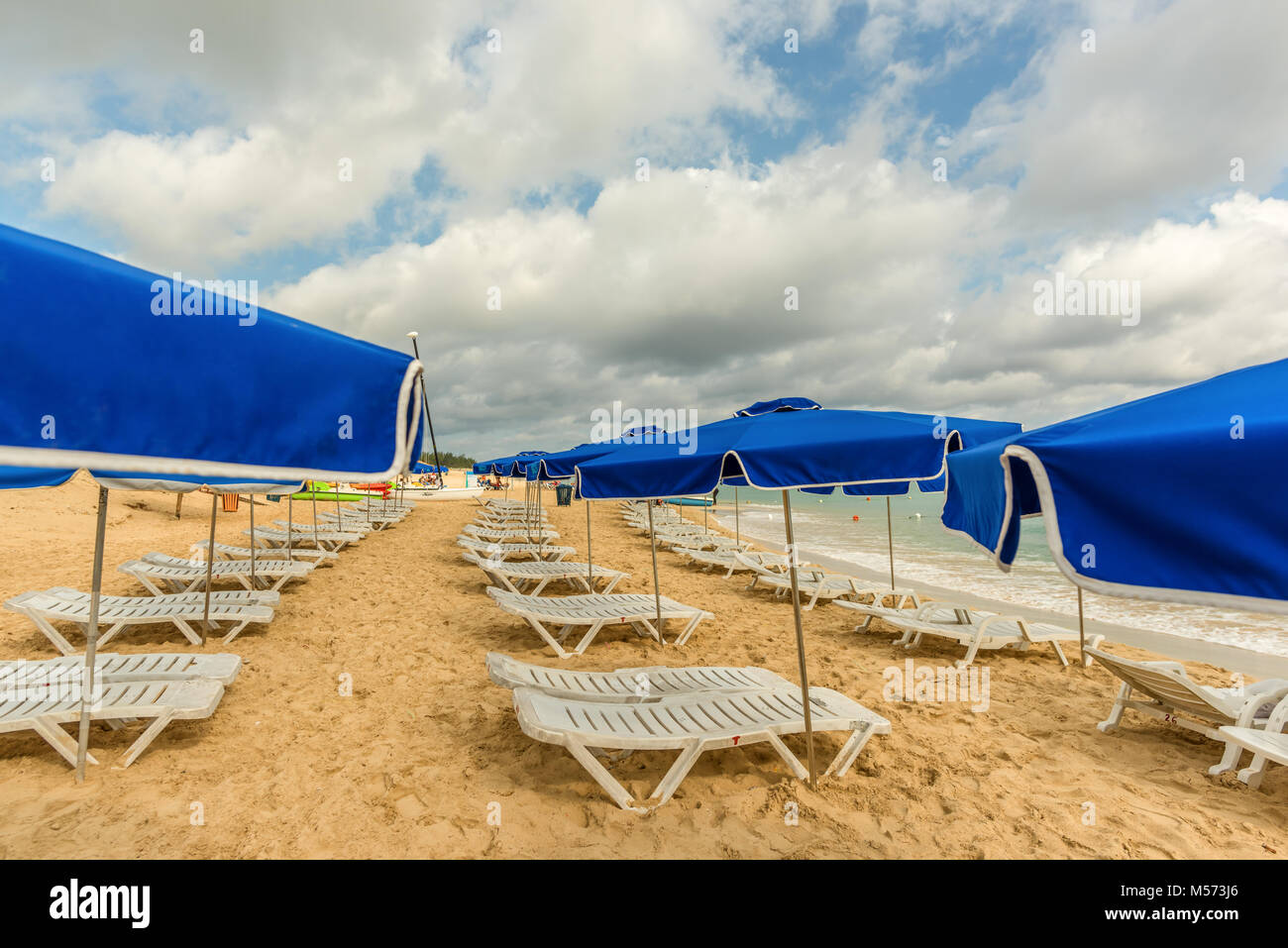 White sdraio e ombrelloni blu stand su una spiaggia di sabbia vicino al mare con onde verdi e un cielo blu con nuvole bianche Foto Stock