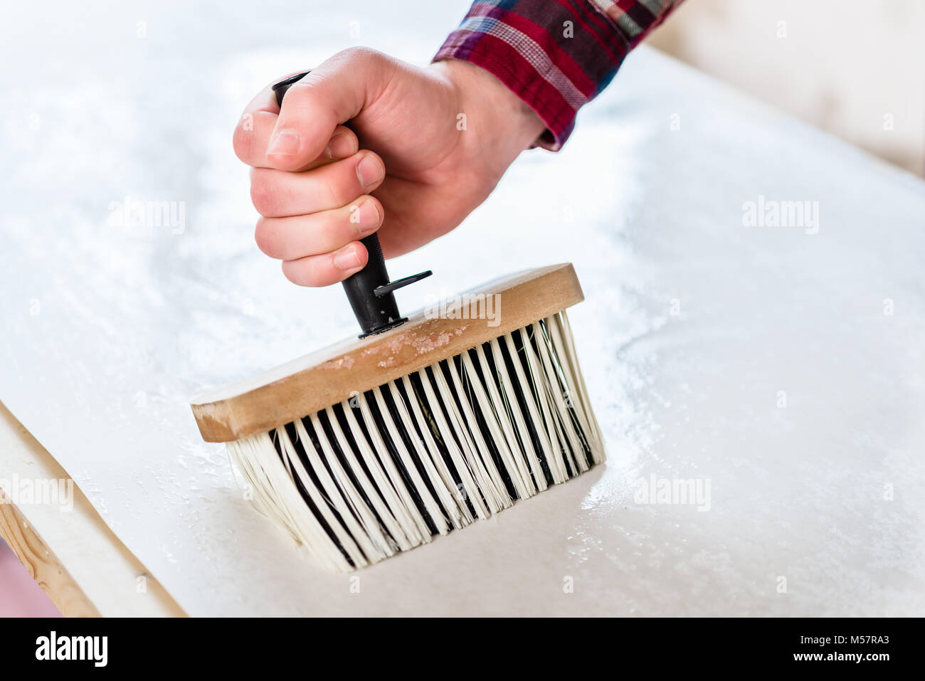 Close-up della mano di un uomo con una spazzola sintetica con manico in plastica durante la applicazione di pasta alla superficie del foglio di carta da parati Foto Stock