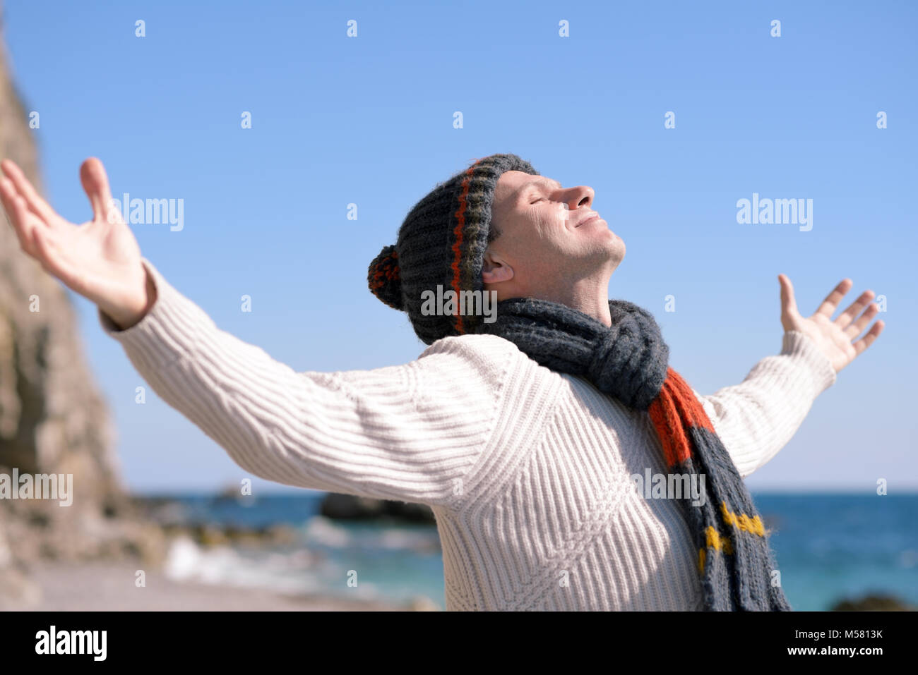 Uomo felice nel cappello di maglia e sciarpa su una spiaggia sotto i raggi del sole Foto Stock