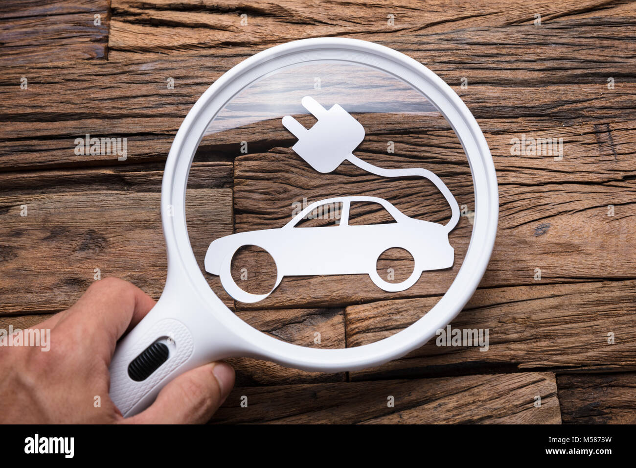 Immagine ritagliata di imprenditore il mano che tiene la lente di ingrandimento su auto elettriche al tavolo di legno Foto Stock