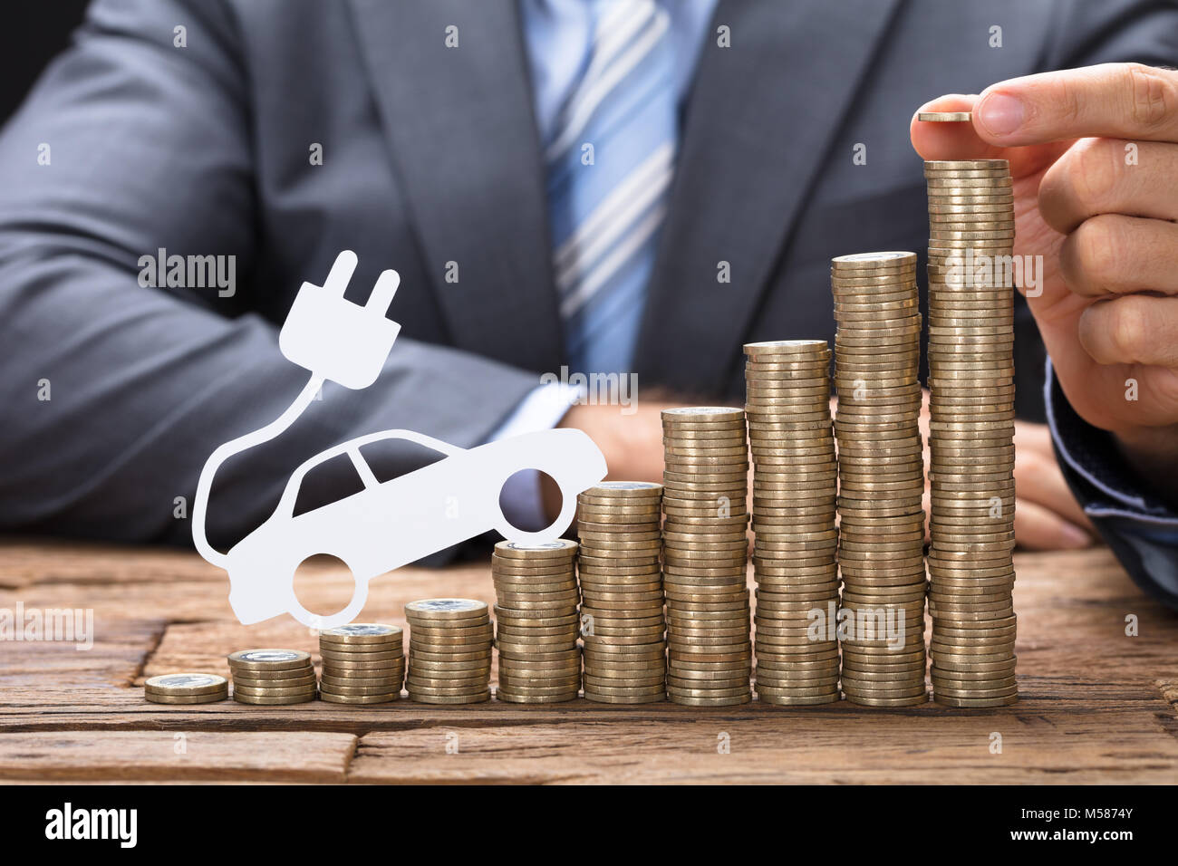 Sezione mediana di imprenditore di monete di impilamento con auto elettrica al tavolo di legno Foto Stock