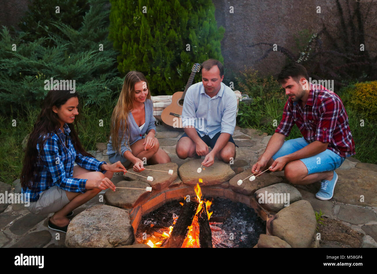 Compagnia di giovani azienda bastoni con marshmallows al di sopra del fuoco di sera. Vacanza e amicizia concep Foto Stock