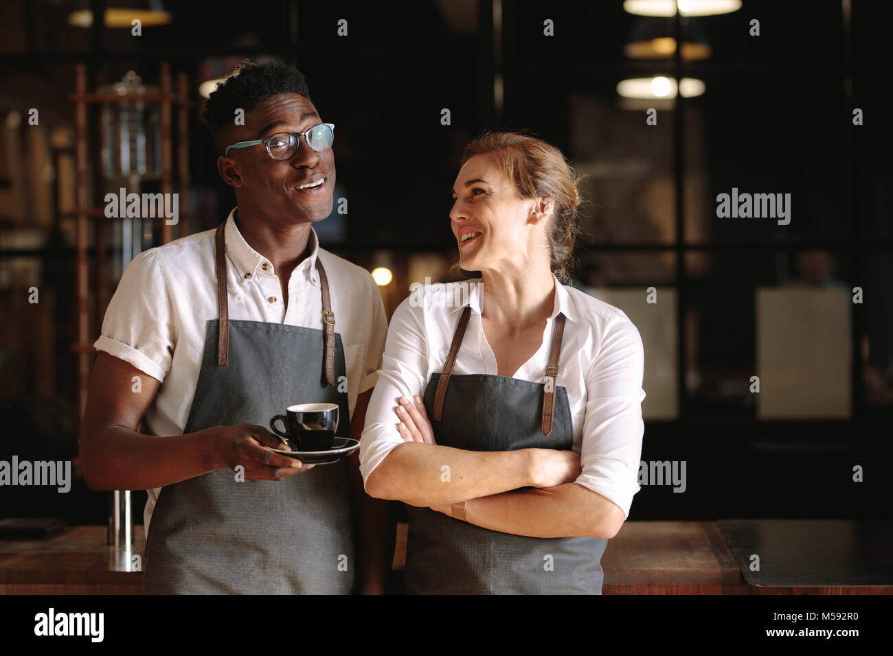 Caffè felice proprietario di un negozio di giovane in piedi all'interno del loro negozio. L uomo e la donna baristi in piedi all'interno del loro café indossando il grembiule. Foto Stock