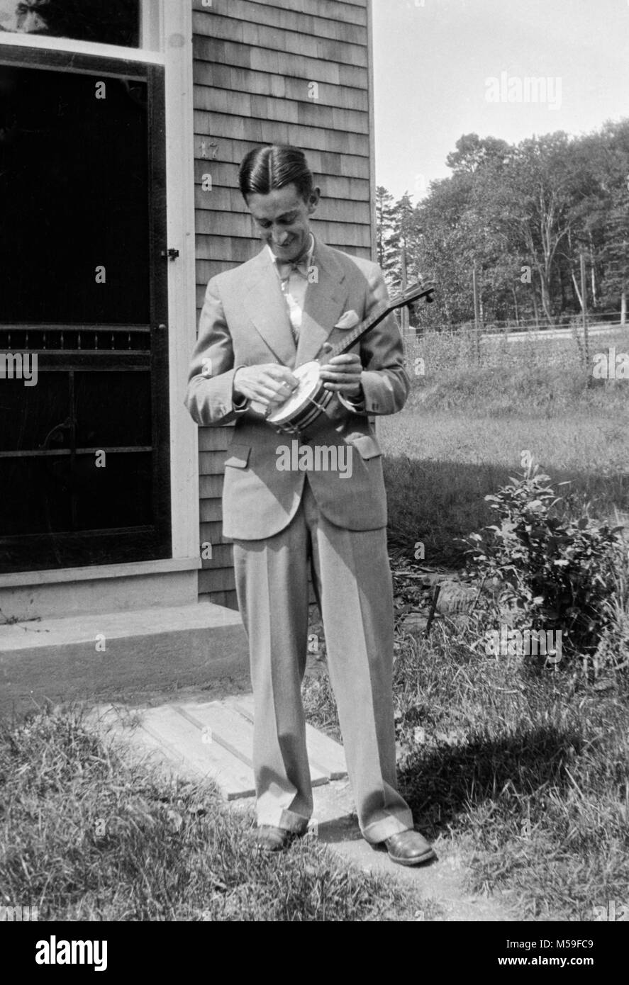 Un giovane uomo spenna a banjo tenore al di fuori di casa sua, ca. 1930. Foto Stock