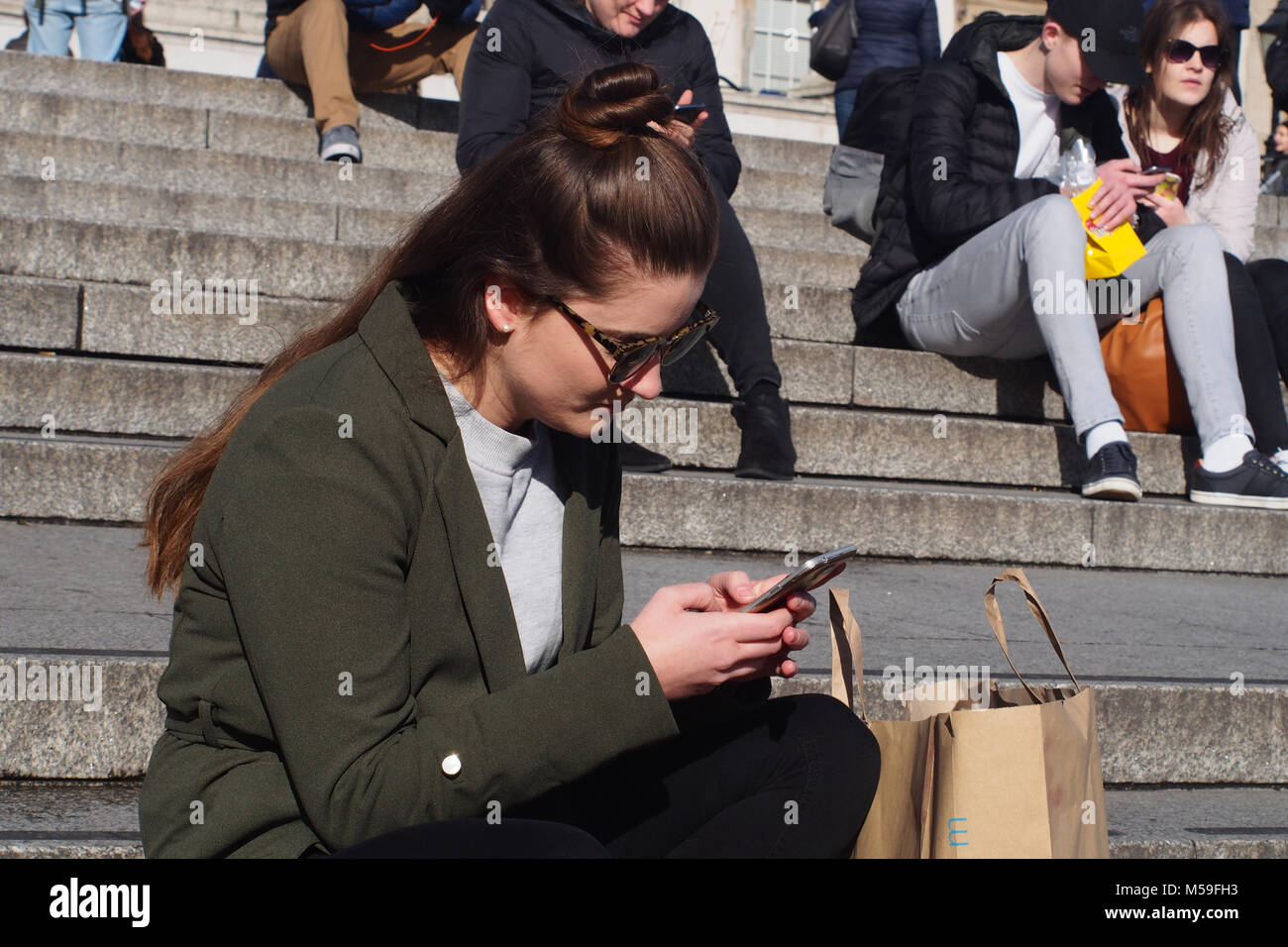 Una giovane donna seduta sui gradini di pietra guardando il suo smartphone in inverno il sole, Trafalgar Square, Londra Foto Stock