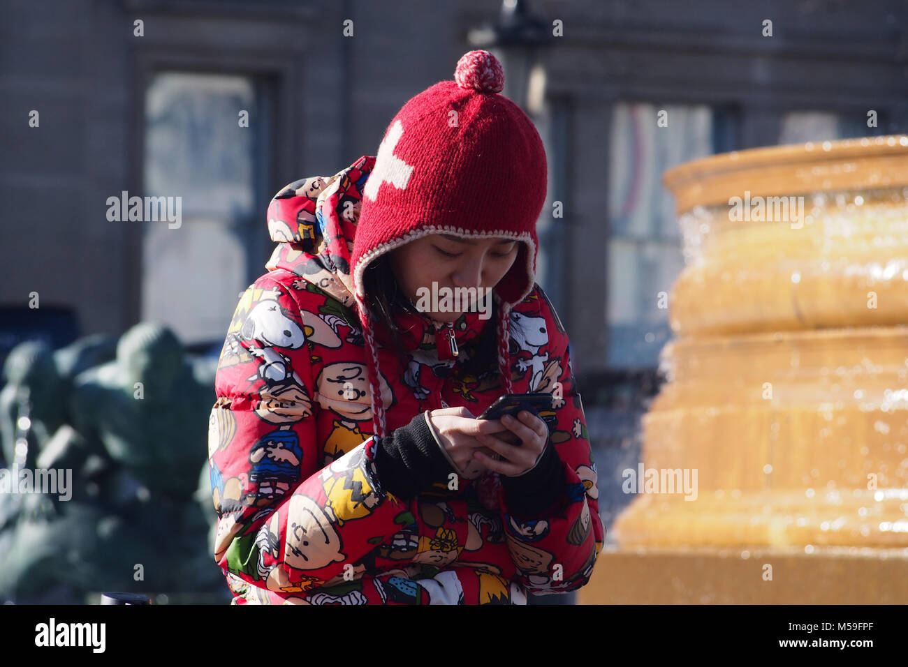 Una giovane donna orientale con il suo smartphone in strada in un freddo luminosa giornata invernale e caldo da indossare abbigliamento invernale, Trafalgar Square, Londra Foto Stock