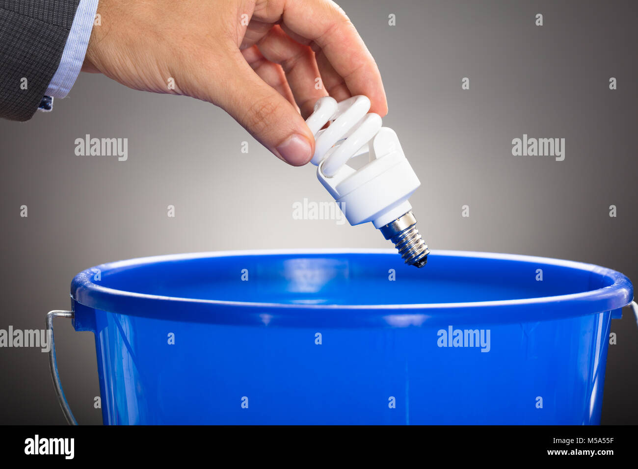 Ritagliate la mano dell'uomo d'affari gettando efficiente dal punto di vista energetico lampadina in blu la benna contro uno sfondo grigio Foto Stock