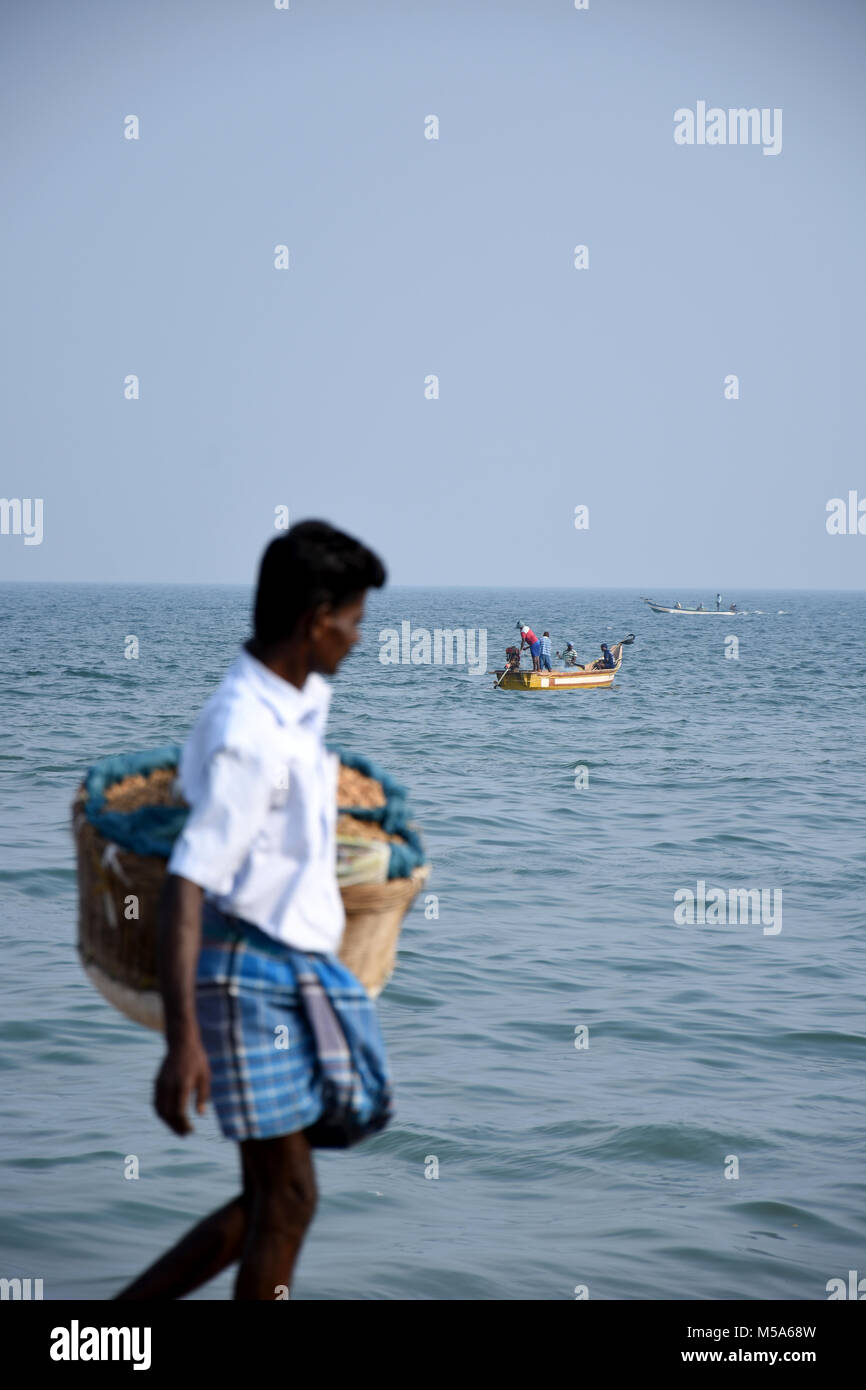 Gli uomini di Fisher impostazione delle sue reti in mare e un occupato arachide venditore passando sulla spiaggia. Foto Stock