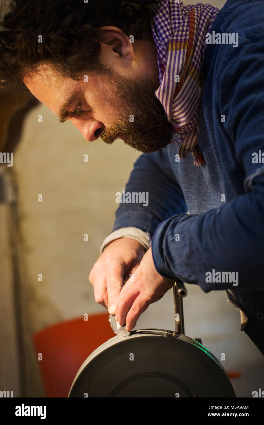 Close up di un artigiano in un workshop, tenendo la lama in acciaio di un coltello su un asse rotante ruota di levigatura o smerigliatrice. Foto Stock
