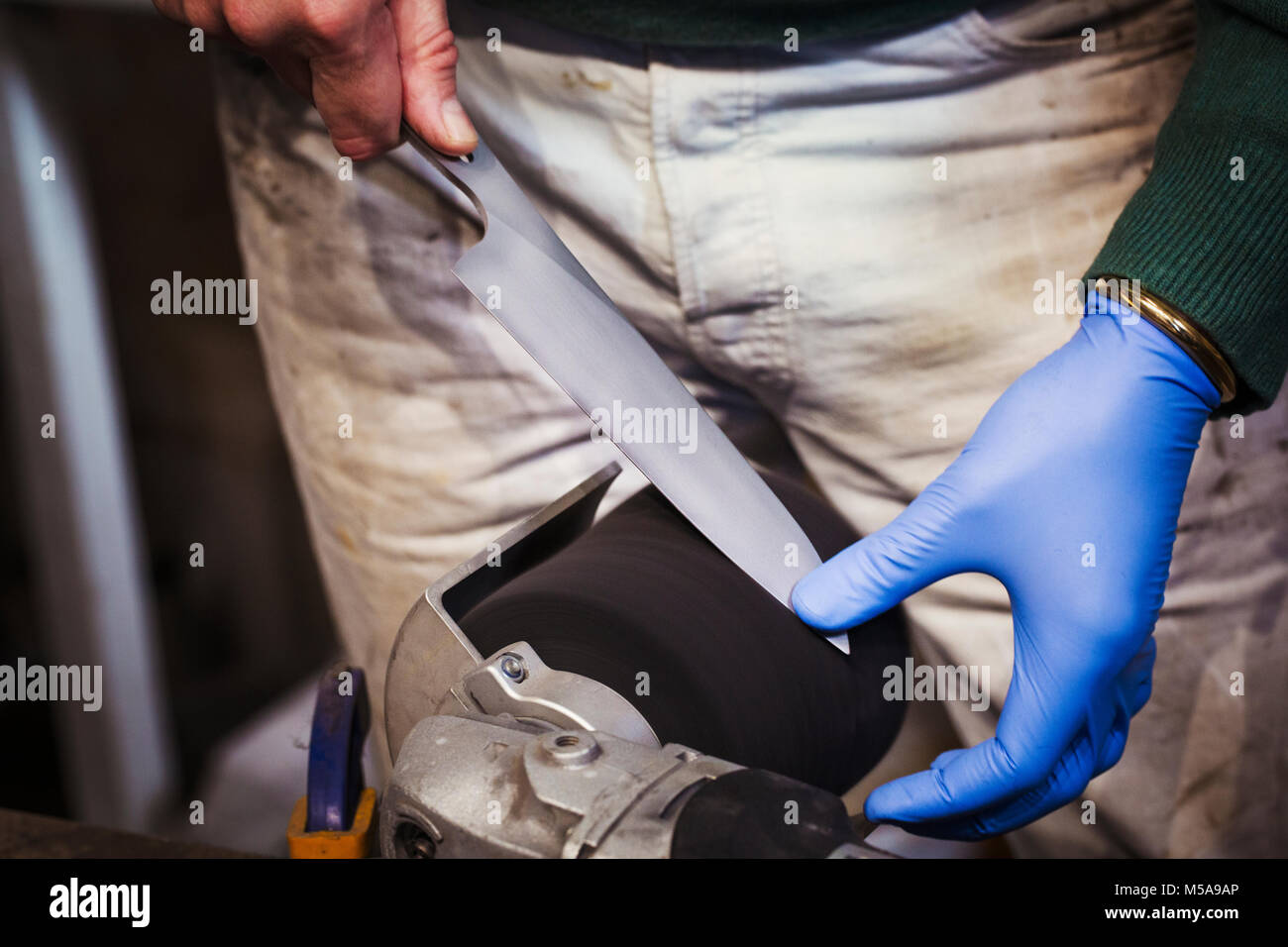 Close up di un artigiano la mano in un guanto blu, affilatura e la levigatura della punta di un coltello lama su una mola. Foto Stock