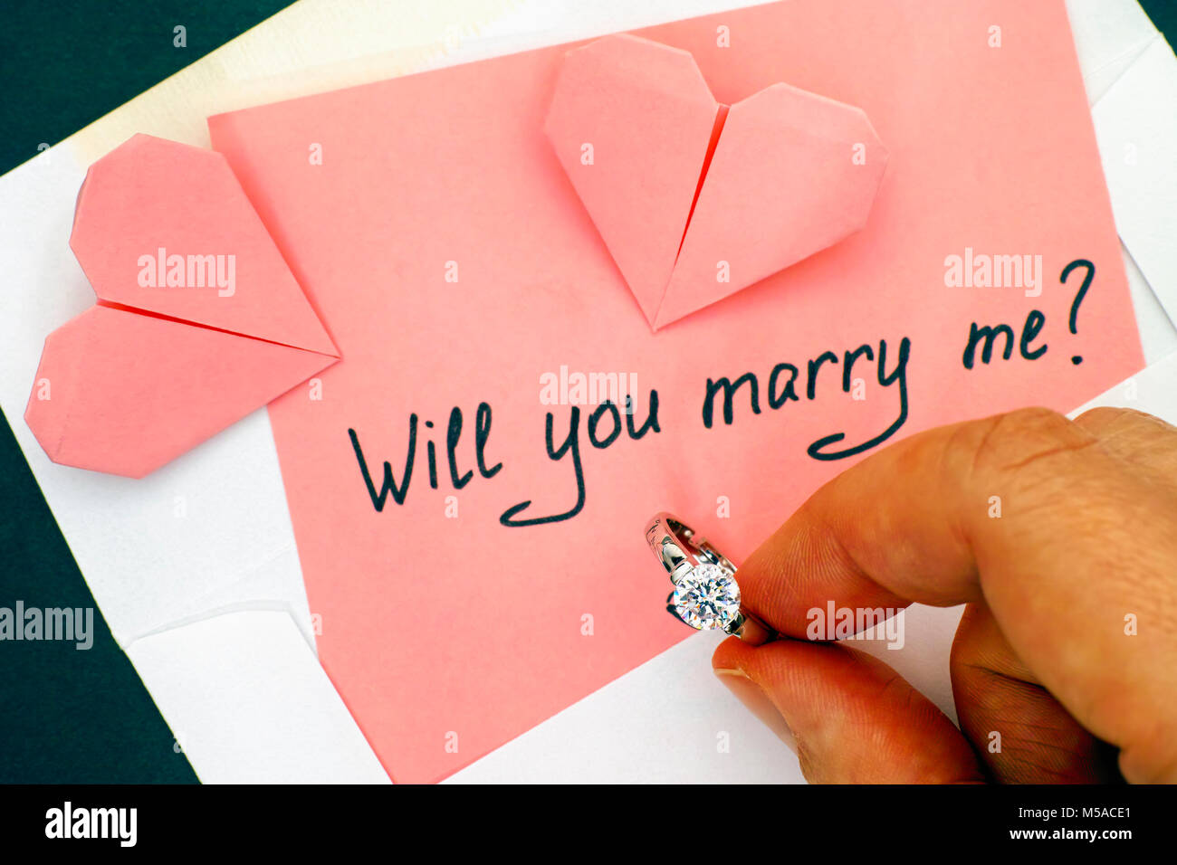 La persona tenuta a mano anello di fidanzamento. Lettera con testo ti sposerà me? In busta e due cuori origami sul tavolo. Foto Stock