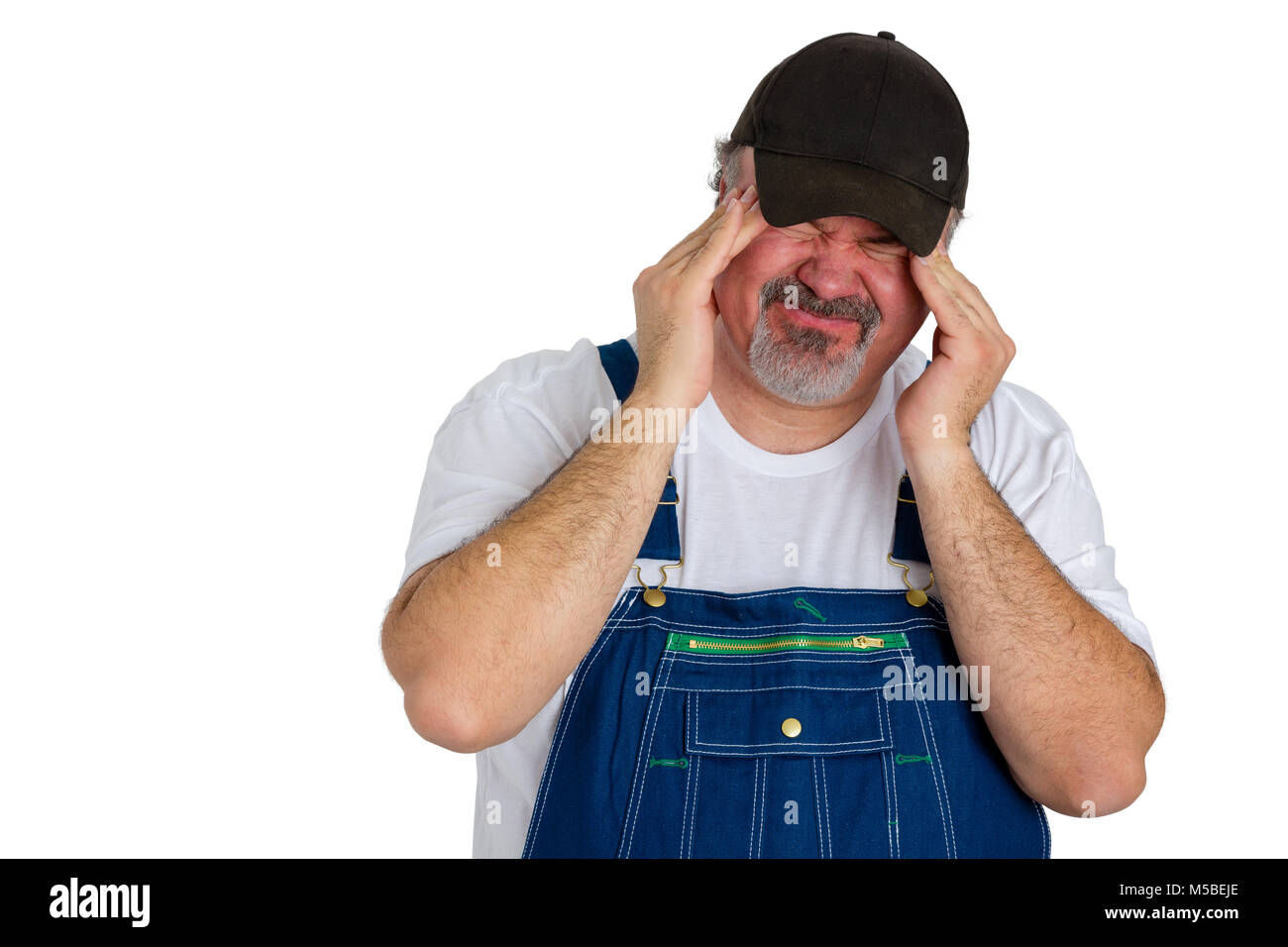 Workman in bib tute con un mal di testa di emicrania smorfie con il dolore acuto come egli sfrega contro i suoi templi con i polpastrelli delle dita in un sanitario e medico Foto Stock