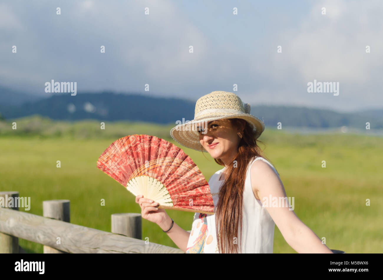 Capelli lunghi, appoggiata contro il corrimano in legno, una camicia bianca, una felice giovane donna. Vi è una ventola in mano. Su una primavera piovosa giornata, Foto Stock