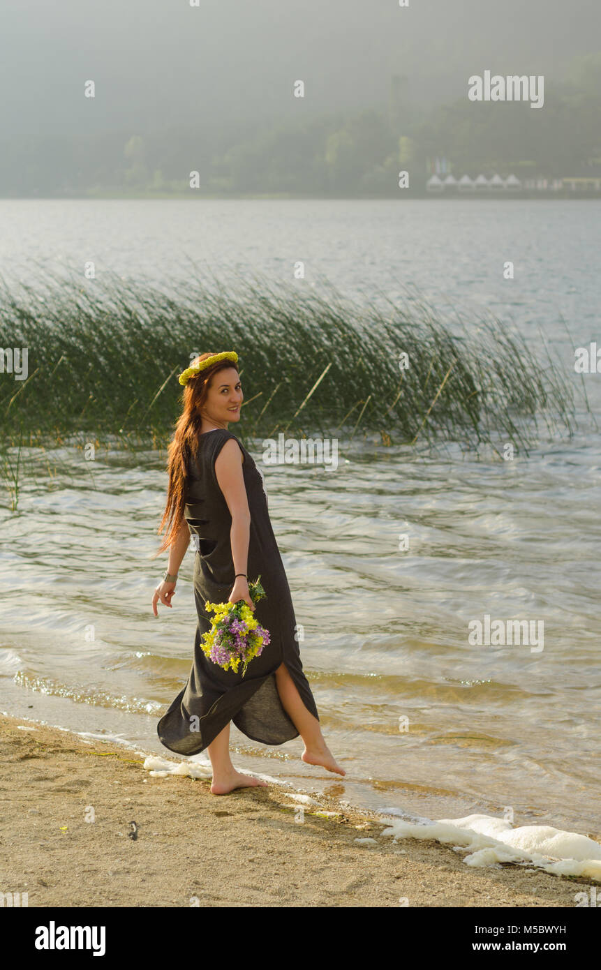 Giovane Donna con fiori di colore giallo nella sua mano in riva al lago, lungo abito nero, con il suo piede destro esteso per l'acqua. Foto Stock