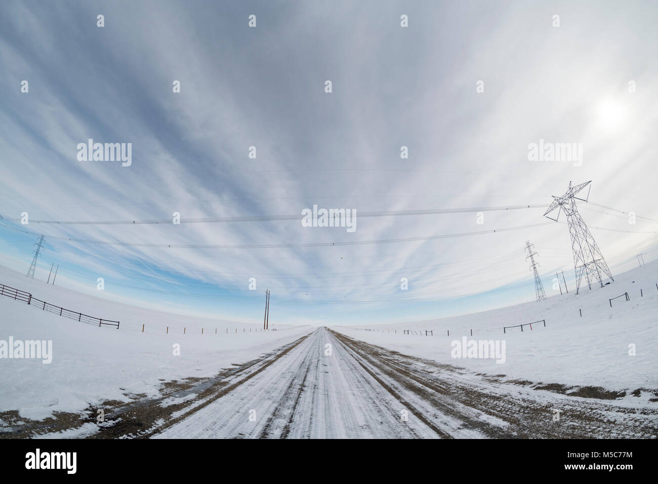 Alberta, Canada. Linee di trasmissione di potenza attraversando una strada di ghiaia sulle praterie in inverno, fisheye prospettiva. Foto Stock