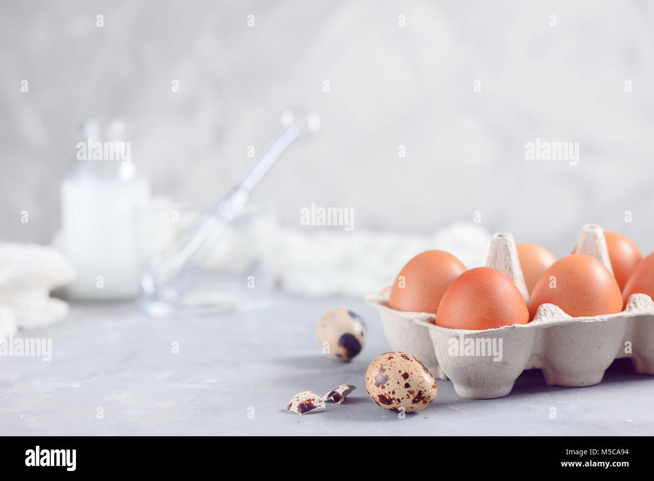 Foglio di carta cartone di uova di colore marrone su sfondo chiaro con uova di quaglia, frusta e gli ingredienti per la cottura di Pasqua. Elevato La chiave dello sfondo con copia spazio. Foto Stock