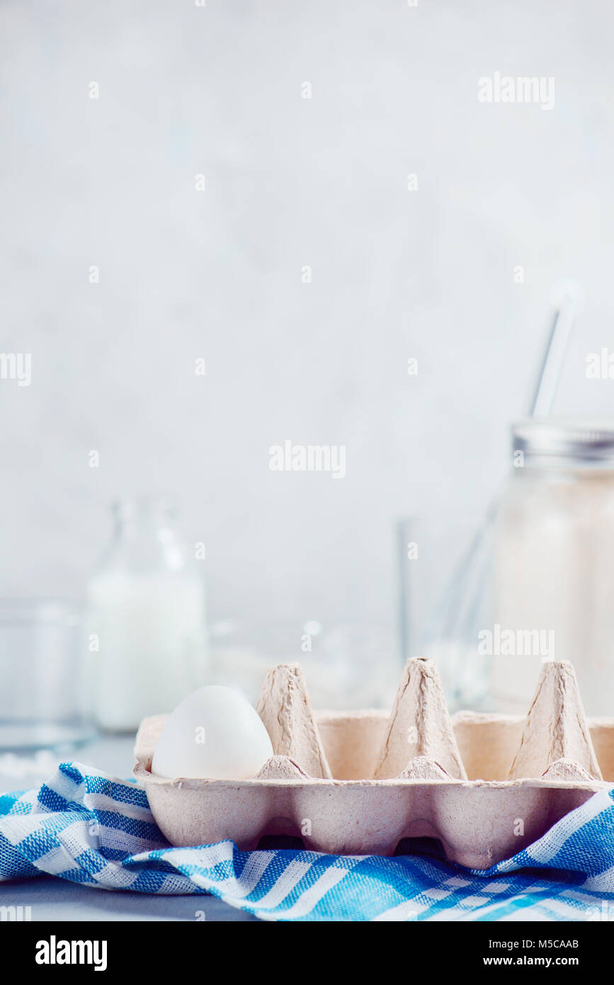 Solitaria uovo bianco in un vassoio della carta su uno sfondo luminoso con il latte, la farina e gli ingredienti per la cottura di Pasqua. Elevato La chiave dello sfondo con copia spazio. Foto Stock