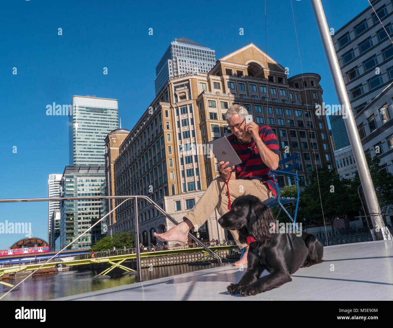 Uomo maturo city location & spaniel cane fuori su office barge boat parlando su smartphone iPhone & iPad controllo computer Canary Wharf London E14 Foto Stock