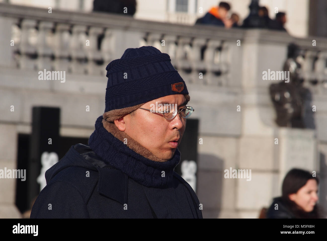 La testa e le spalle di un uomo orientale in Trafalgar Square, Londra, indossando un cappellino, sciarpa e inverno del mantello e in inverno il sole Foto Stock