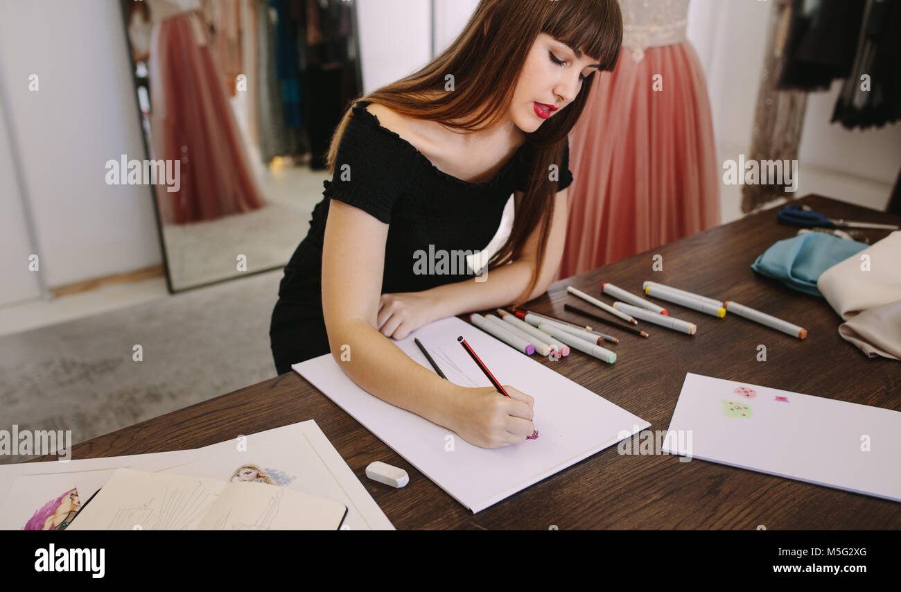 Moda femminile imprenditore abbozzare un disegno seduto alla sua tavola. Designer di moda fare un disegno nel suo negozio di stoffa. Foto Stock