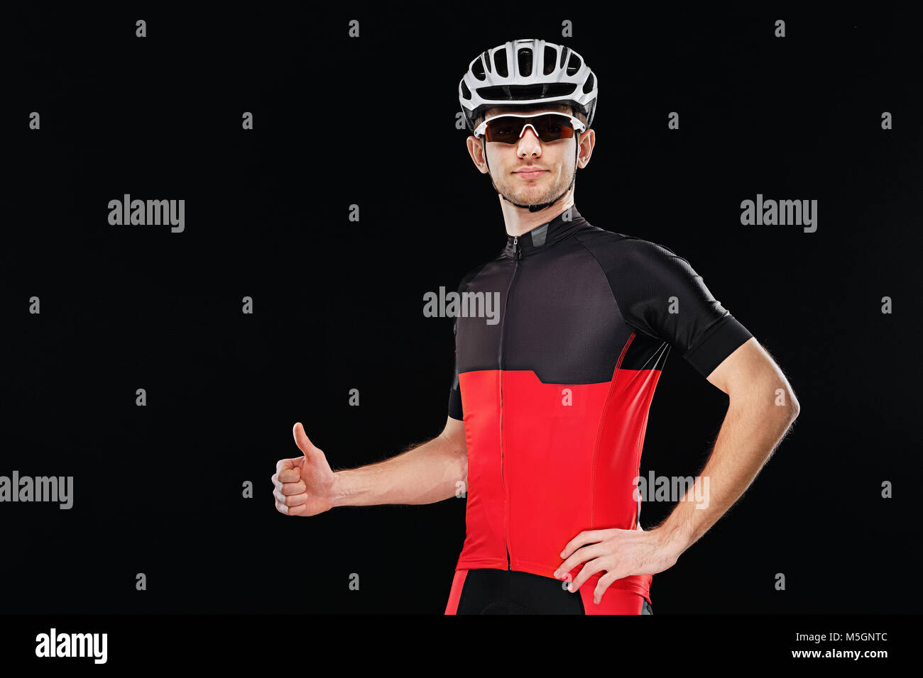 Sport. Ciclista nella formazione di vestiti, occhiali da sole e casco mostrando pollice in alto segno su sfondo nero. Foto Stock