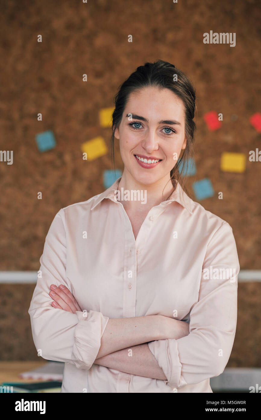Ritratto di una femmina di lavoratore di ufficio. Lei è in piedi di fronte ad una bacheca di sughero, elegantemente vestito con le braccia piegate e sorride per la fotocamera. Foto Stock