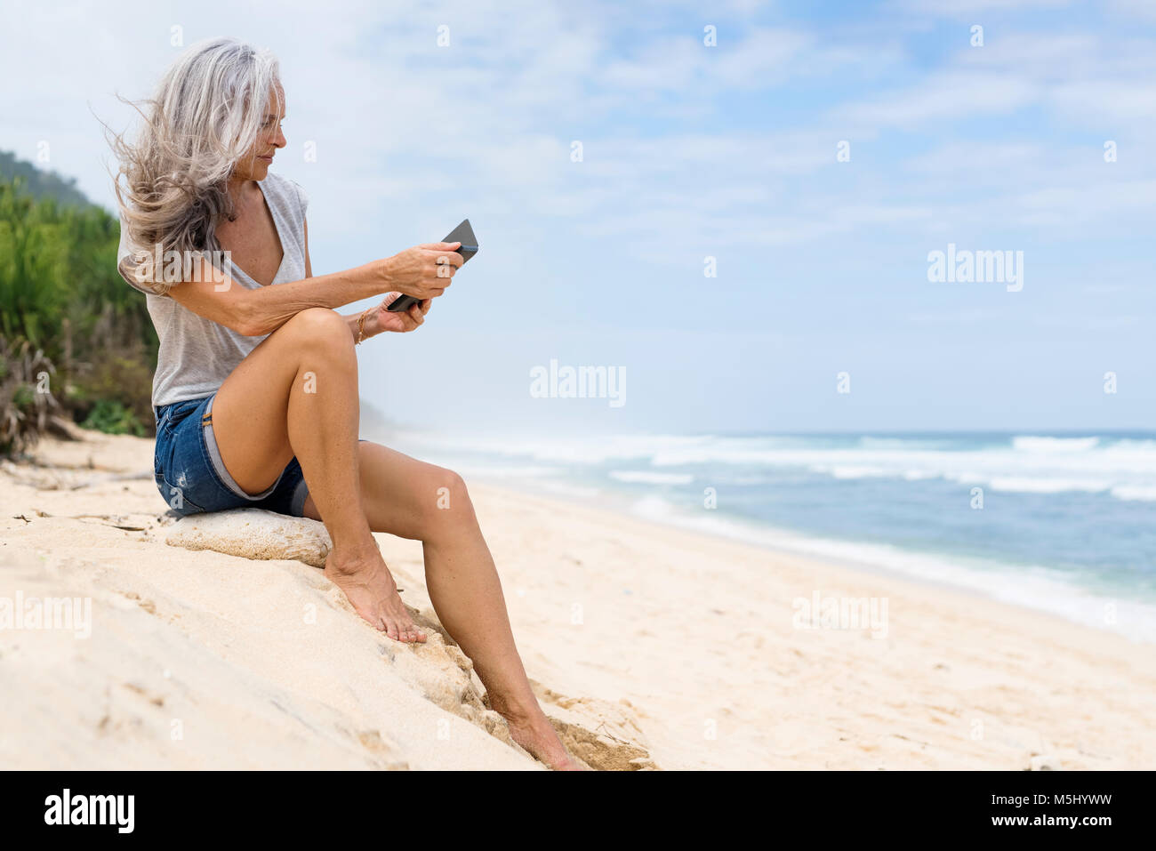 Bella sorridente senior donna seduta sulla spiaggia, azienda e-book Foto Stock