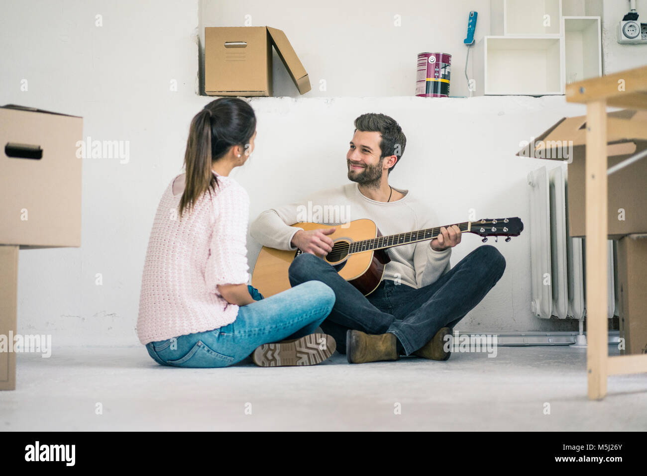 Giovane seduto sul pavimento nella nuova casa a suonare la chitarra Foto Stock