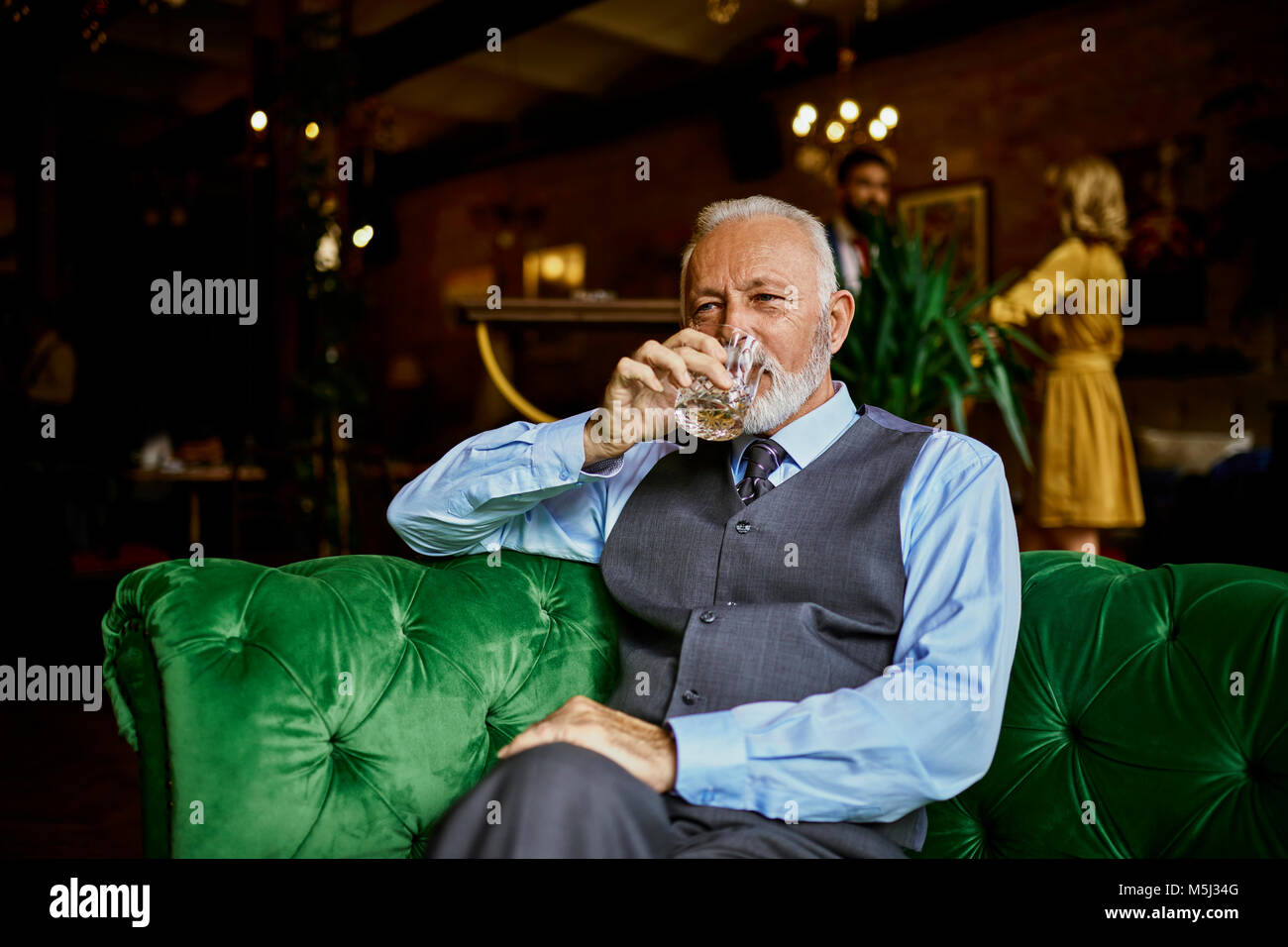 Ritratto di senior elegante uomo seduto sul lettino in un bar a bere dal bicchiere Foto Stock