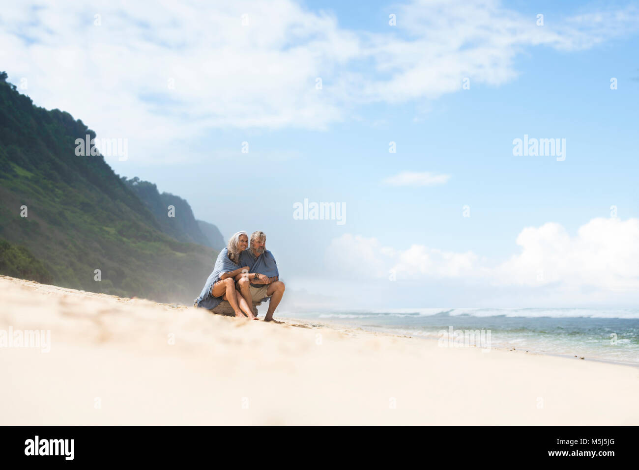 Coppia senior seduto sulla spiaggia, avvolto in un mantello Foto Stock