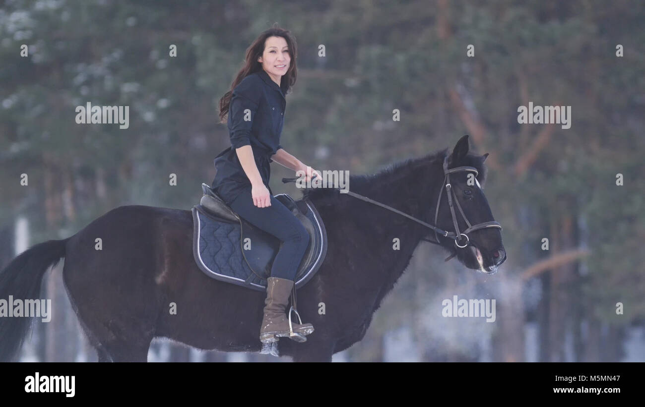 Bella donna longhaired in sella ad un cavallo nero attraverso la neve nella foresta Foto Stock Bella donna longhaired in sella ad un cavallo nero attraverso la neve nella foresta Foto Stock