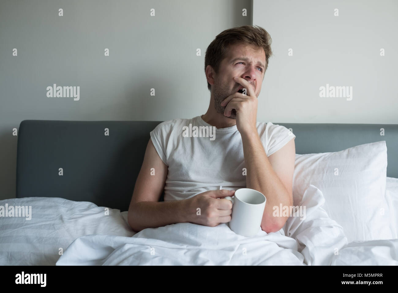 Giovane uomo bello sbadigliare e avente il caffè del mattino a letto Foto Stock