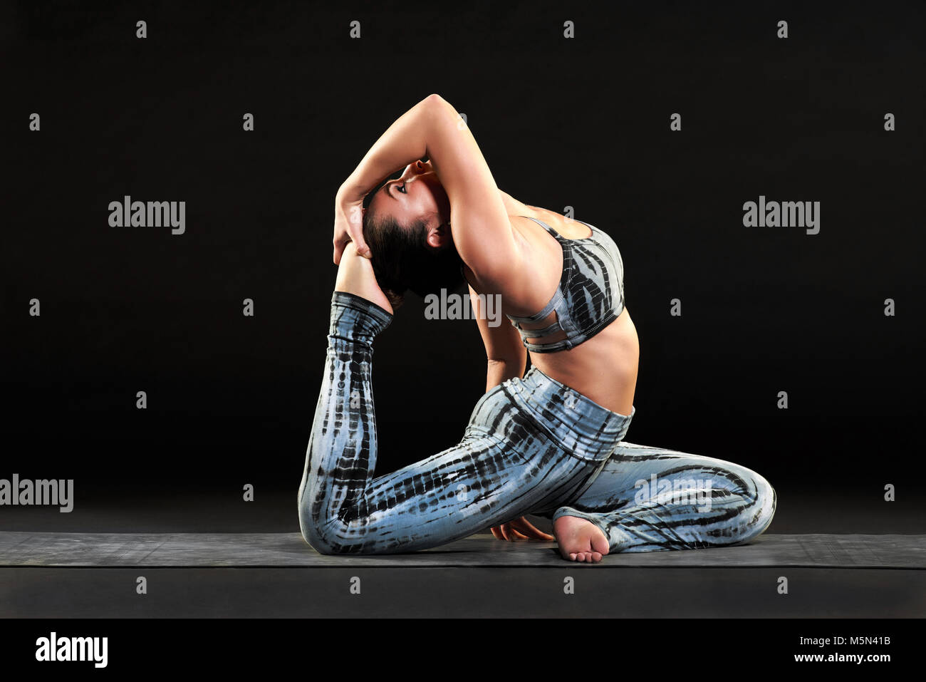 Montare morbida donna facendo un re pigeon yoga pongono inarcamento torna ad abbracciare il suo piede in una vista laterale su uno sfondo nero Foto Stock