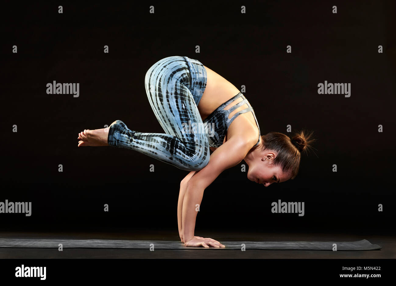 Montare Atletica Giovane donna dimostrando una cornacchia pongono yoga in equilibrio su le braccia con il suo corpo in una posizione arricciata, vista laterale sul nero Foto Stock