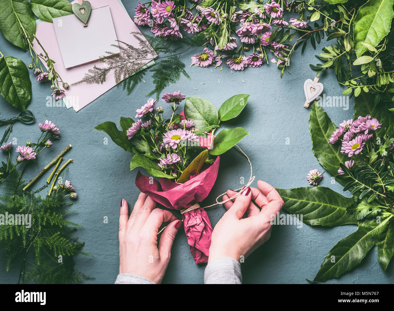 Mani femminili rendendo bouquet di fiori disposizione con foglie verdi di decorazione su lavoro tabella grigia con un fioraio strumenti e accessori, vista dall'alto, fase Foto Stock