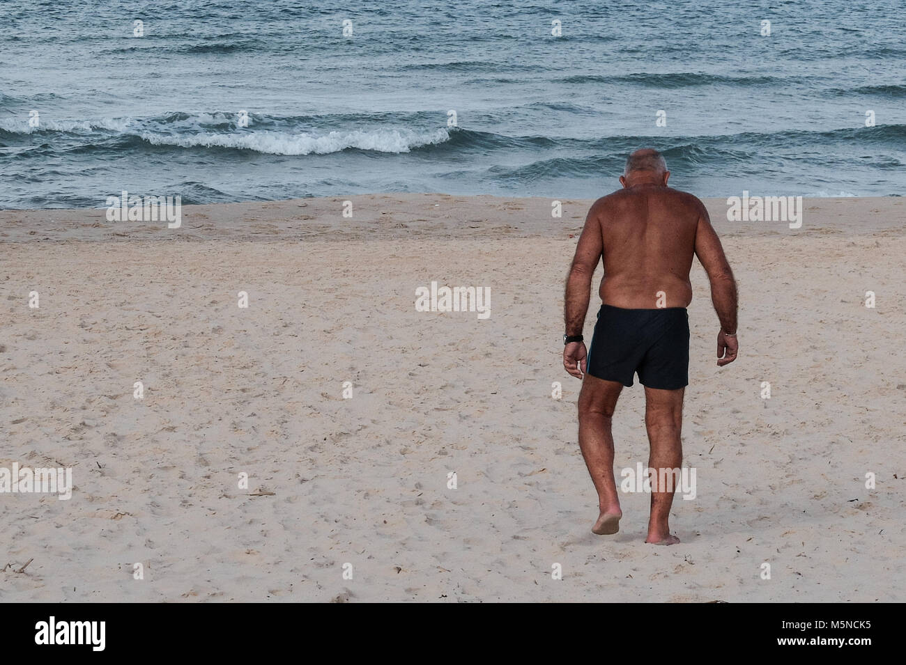Un uomo anziano in un costume da bagno cammina verso l'acqua su di una spiaggia di sabbia. Foto Stock