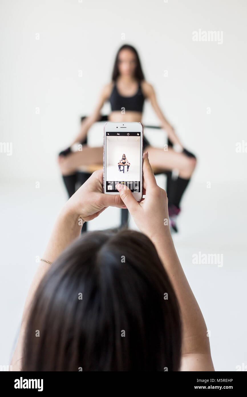 La riflessione nel telefono cellulare. La donna sta prendendo la foto della sua attivitã sportive ragazza in sportwear in posa sulla sedia in bianco lo studio. Foto Stock