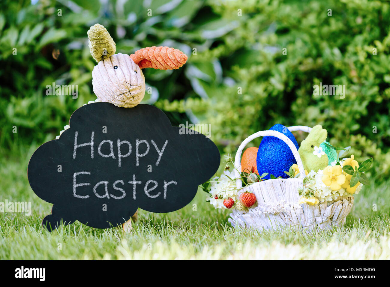 Toy bunny con chalk testo 'Felice Pasqua' sulla lavagna e cesto con uova, fiori e frutti di bosco Foto Stock