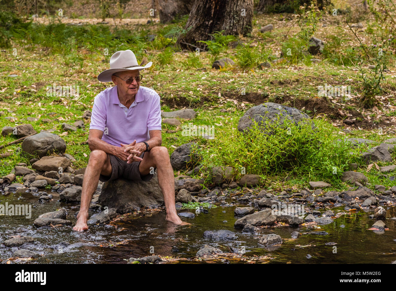 Anziani uomo Australiano, indossando un stetson, seduta da un torrente in alto la Hunter Valley, NSW, Australia. Foto Stock