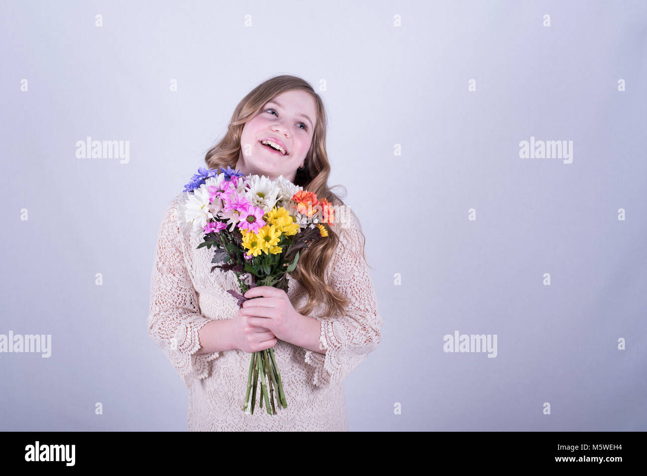 Dodici-anno-vecchia ragazza con lunghi, sporchi capelli biondi holding colorato bouquet di margherite sorridenti, guardando verso l'alto e di lato, sfondo bianco Foto Stock