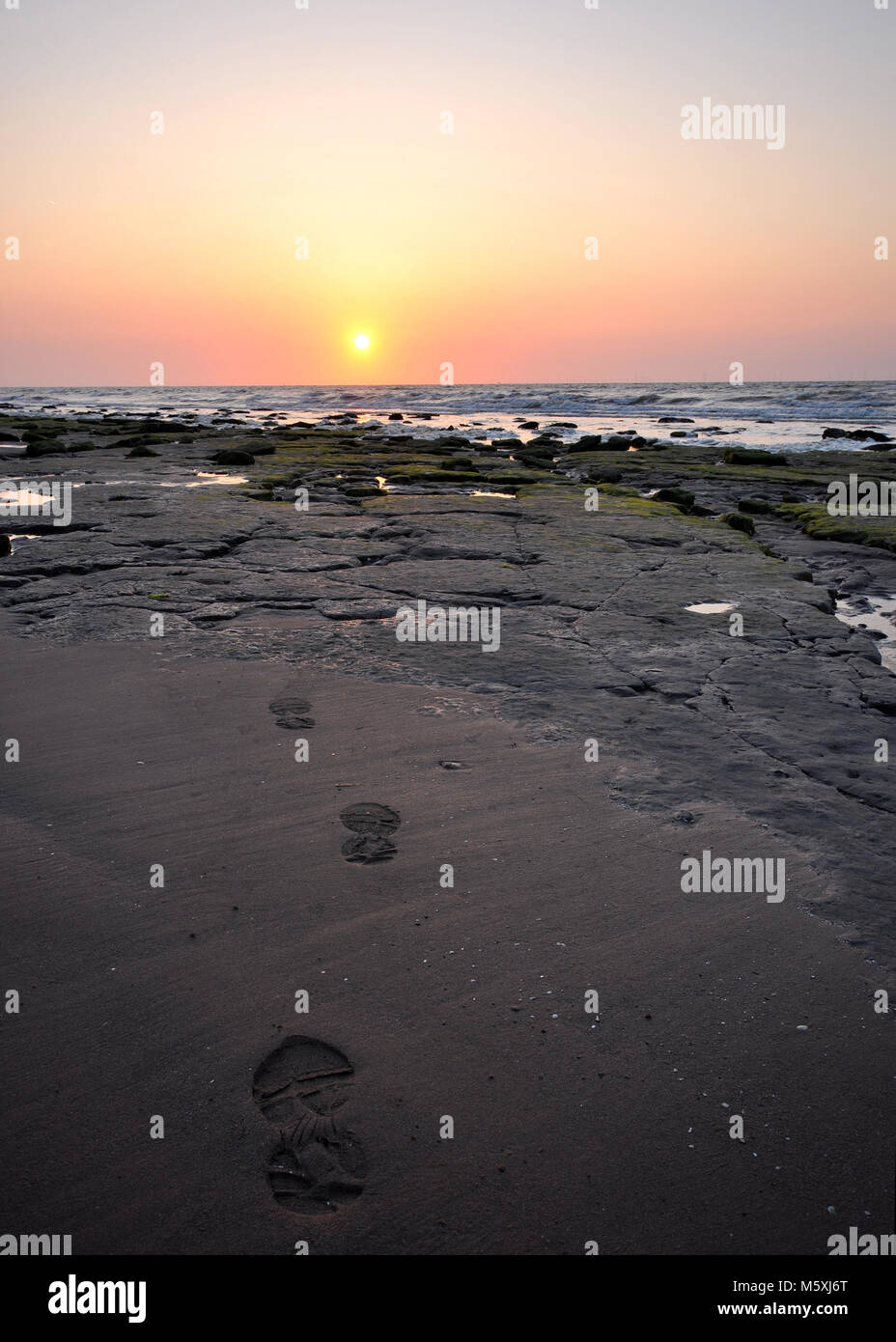 Foto della spiaggia con sabbia, rocce e impronte che conducono ad un mare al tramonto a Reculver Beach, Kent Foto Stock