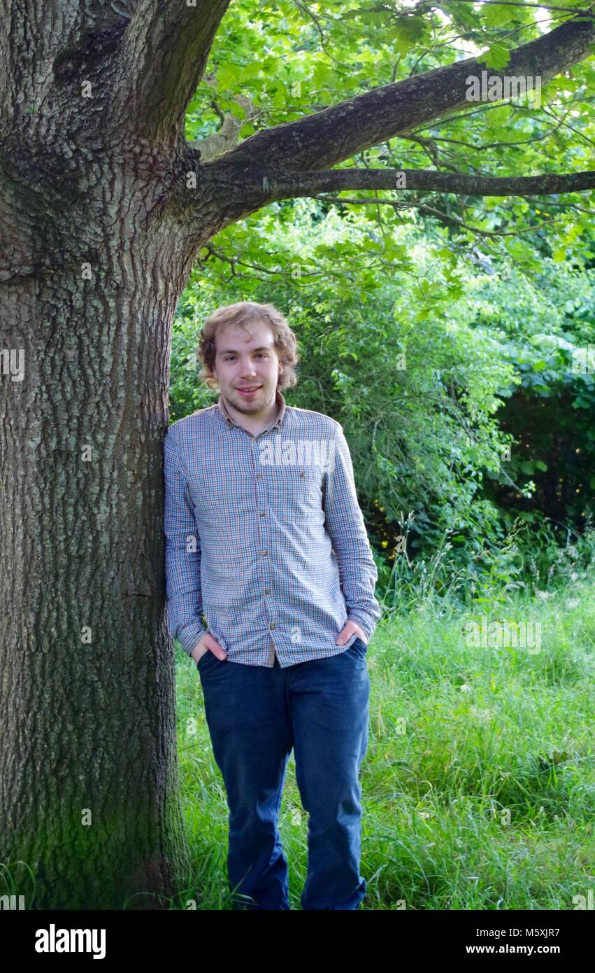 Giovane uomo bello, appoggiata contro un inglese di quercia in una passeggiata nella natura. Ludwell Valley Park, Exeter, Devon, Regno Unito. Foto Stock